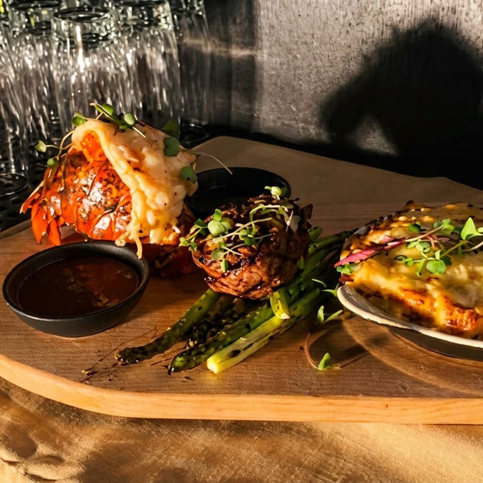 Mill River's surf and turf with lobster, steak, asparagus, and cheesy gratin on a wooden board.
