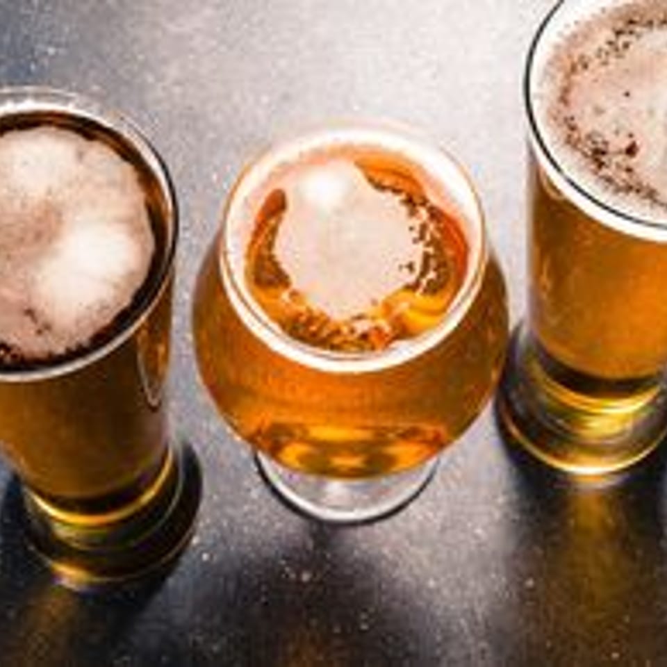 Five glasses of golden beer with foamy heads on a dark table, viewed from above.