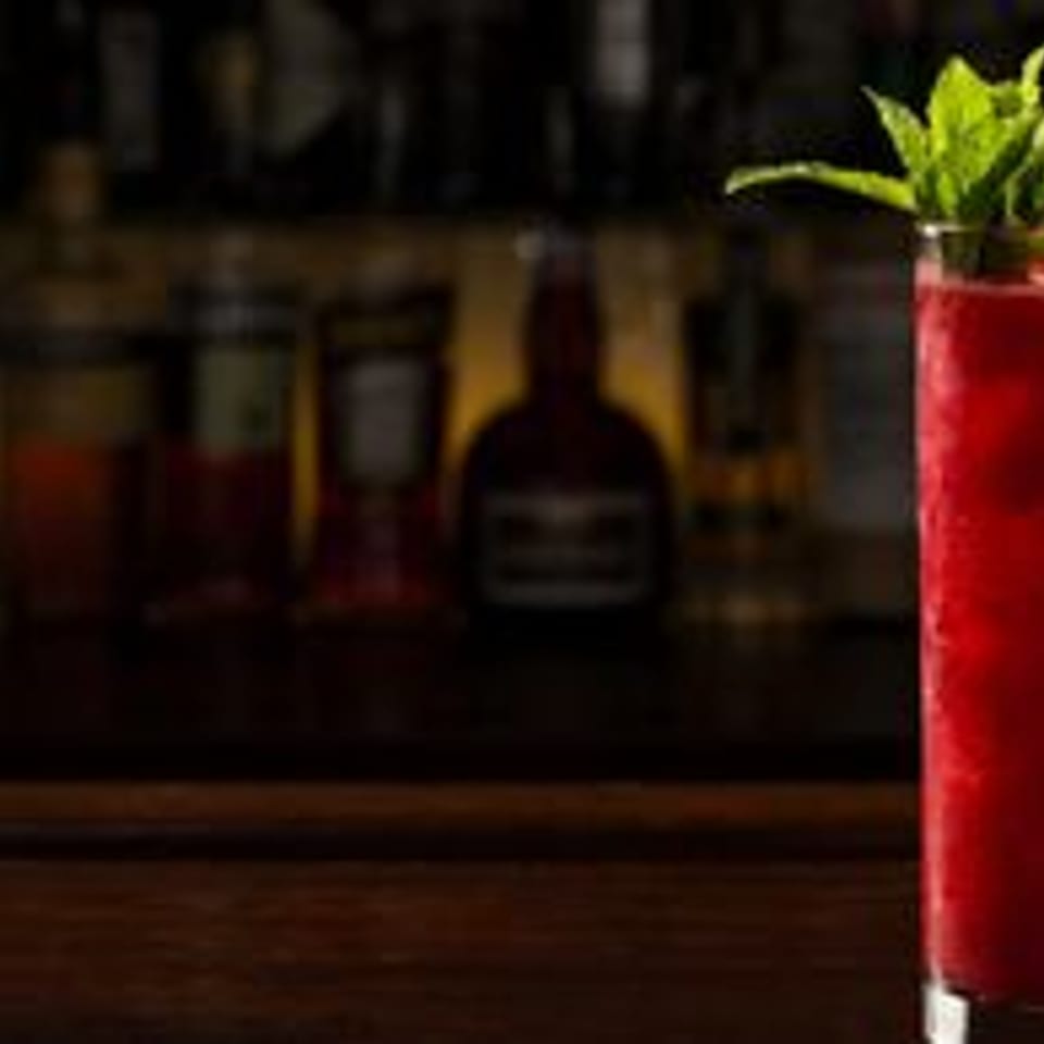 Vibrant red cocktail garnished with fresh mint and berries on a dark bar.