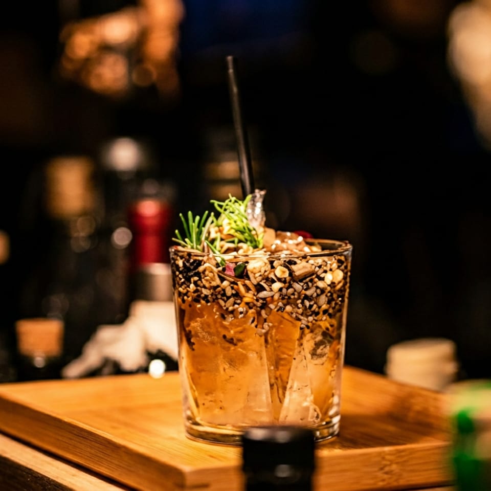 Cocktail with seeds, nuts, and rosemary garnish on a wooden board in a dark bar setting.