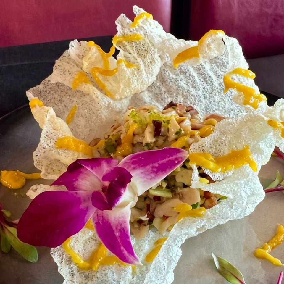 Diced food in a crispy white shell, garnished with a vibrant purple orchid and yellow sauce on a dark plate.