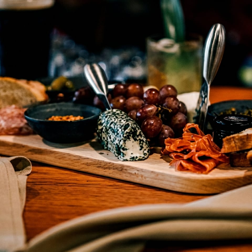 Appetizer board with cheese, grapes, cured meats, bread, and condiments on a wooden table.