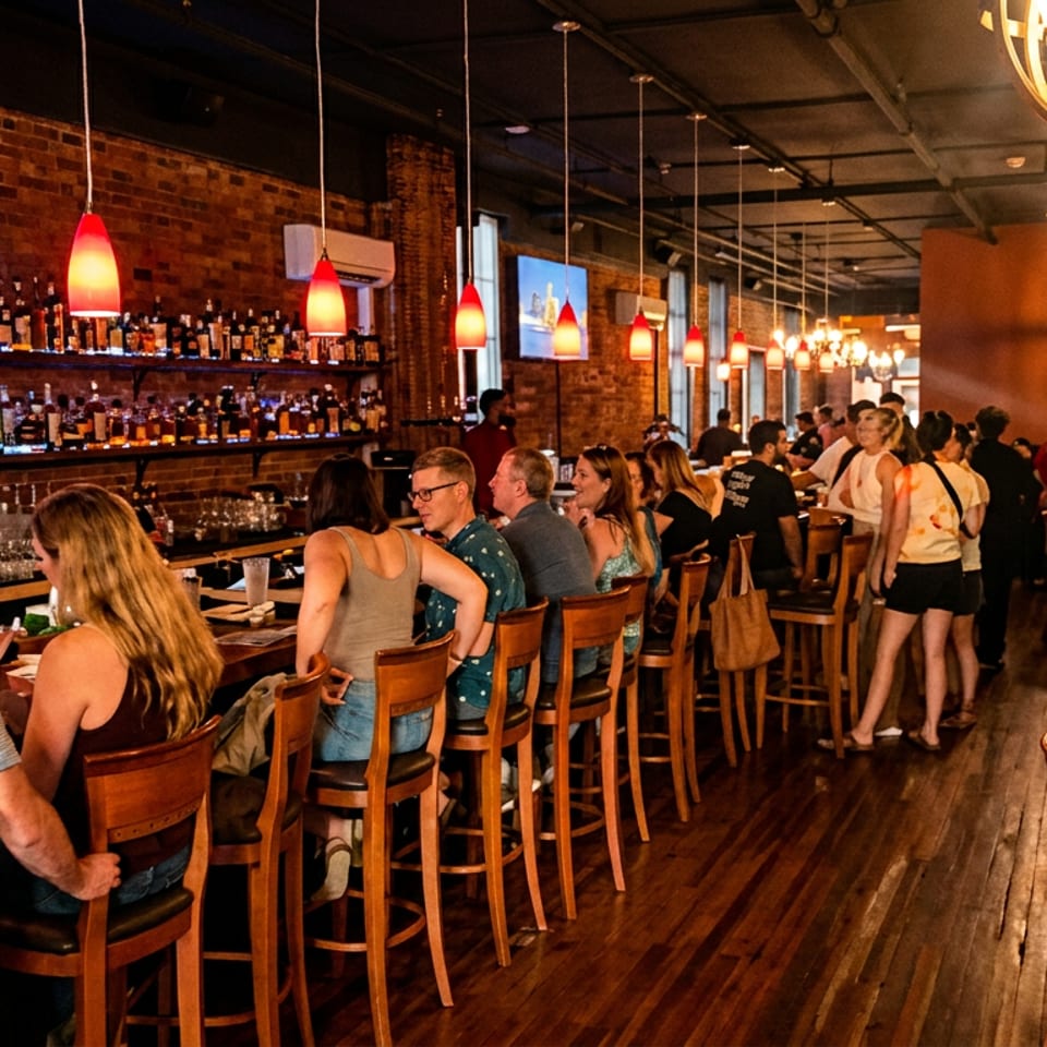 Busy restaurant interior with patrons at a long bar and dining tables, exposed brick walls, and warm lighting.