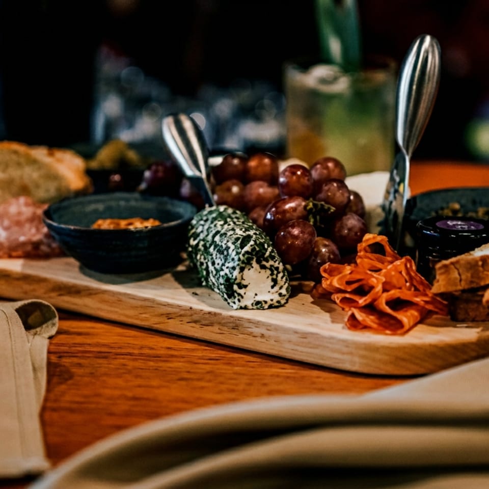 Charcuterie board with herb-crusted cheese, grapes, salami, bread, and dips on a dark wooden table.